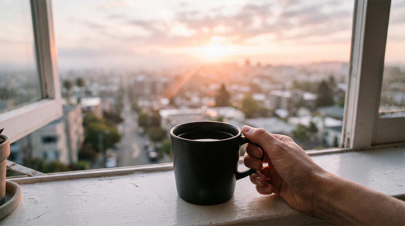A photograph taken from a candid, point-of-view (POV) perspective of right hand holding a matte black ceramic coffee mug filled with coffee on a wooden windowsill. Soft, rose-gold sun rays break through a misty dawn sky over a heavily blurred cityscape (bokeh). There is no ring on her finger. The atmosphere is peaceful, hopeful, and relieved. The image has a natural film grain texture and a shallow depth of field, focusing sharply on the hand and mug.
