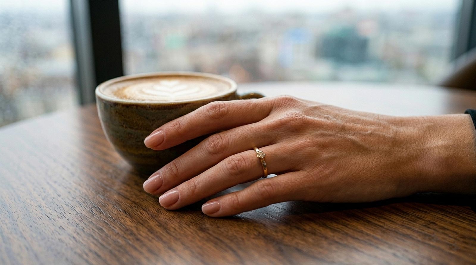 Extreme macro close-up of a woman's hand and a ceramic coffee cup with latte art on a dark walnut table. Natural light highlights textures of skin pores, foam, and wood grain. Symbolic of mindful preparation and intentionality.