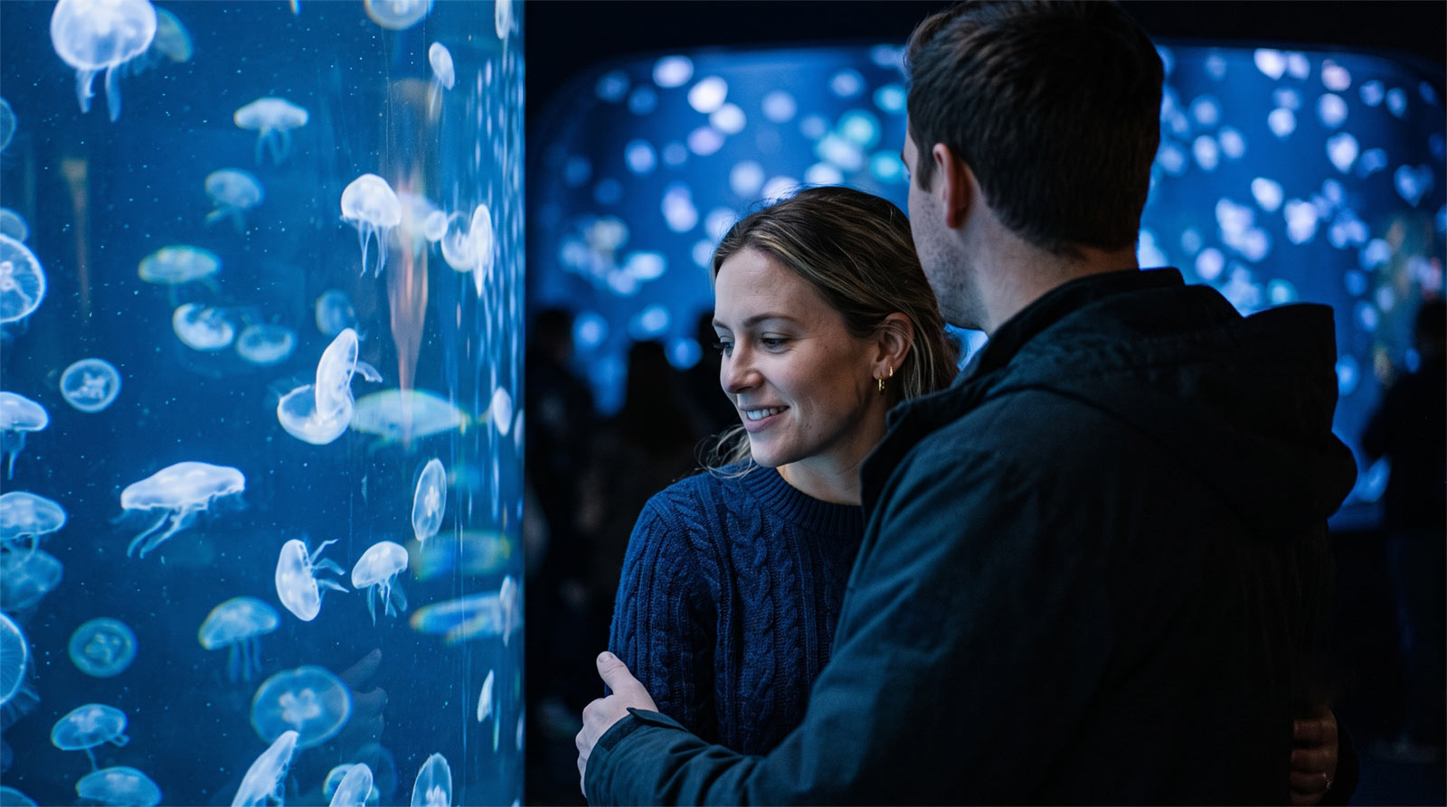 A couple sharing a close, authentic moment illuminated by the ambient blue light of a jellyfish aquarium tank, capturing a modern and intimate date night vibe.